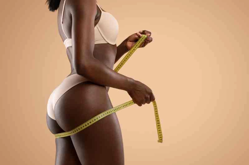 Young skinny african american female in beige underwear with slender body measuring her perfect sexy buttocks with tape measure and showing training result. Studio shot, side view, copy space